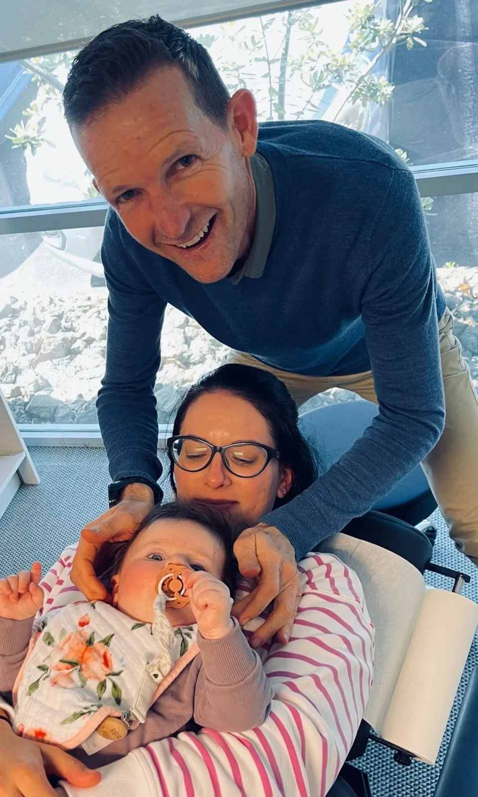 A man and a woman with a baby in a healthcare setting, the man smiling and leaning over, the woman with closed eyes, and the baby with a pacifier, lying on the woman's chest.