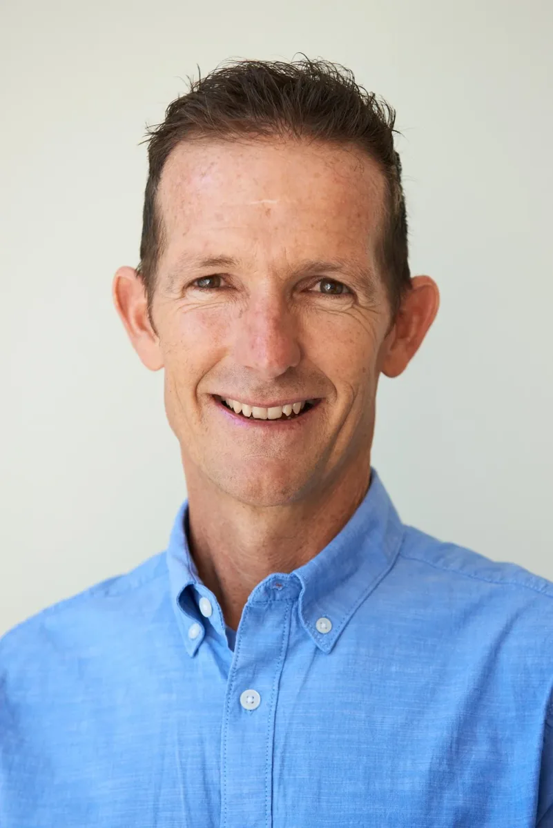 A man with short brown hair wearing a light blue button-up shirt, smiling and looking at the camera.