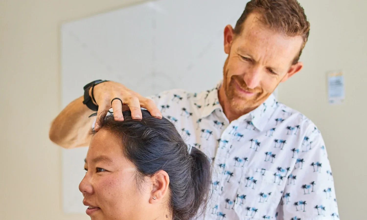 A man with short, curly hair and a beard smiling while gently massaging a woman's head in a clinical setting.